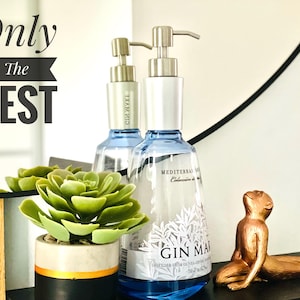 Op de afbeelding: Twee doorzichtige glazen flessen met zilveren pompen, een met het label "Gin Mare" en de andere met "Citrus Grove". De flessen staan op een houten oppervlak met een vetplant in een pot en een gouden aapfiguur.