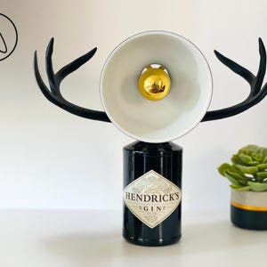 Op de afbeelding: Een unieke lamp gemaakt van een hergebruikte gin-fles, met een witte lampenkap, een gouden lamp en zwarte geweivormige accenten. De lamp is een creatief woondecoratie-item.
