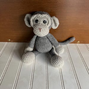May include: A handmade crochet monkey toy crafted from dark gray and light gray yarn. The monkey features a round head with large ears, a light gray muzzle with a stitched nose and mouth, black plastic eyes, a round body, and a long tail.