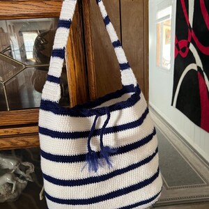 May include: A white and navy blue striped crochet bag with a drawstring closure and a matching strap. The bag is handmade and has a nautical theme. The bag is hanging on a wooden door.