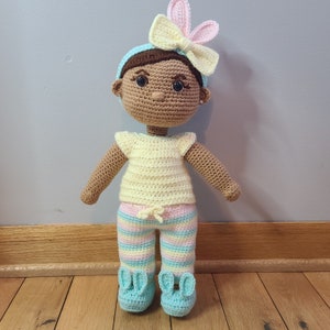 May include: A crocheted doll with brown skin, wearing a yellow and white striped shirt, pink and white striped pants, and blue bunny slippers. The doll has a blue and white headband with a bow.