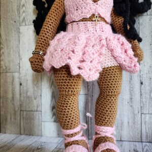 May include: Crocheted doll with brown skin wearing a pink crocheted outfit with a gold ankh pendant. The doll is wearing pink sandals.
