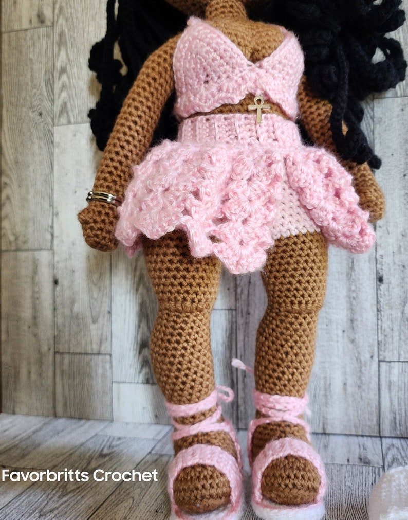 May include: Crocheted doll with brown skin wearing a pink crocheted outfit with a gold ankh pendant. The doll is wearing pink sandals.