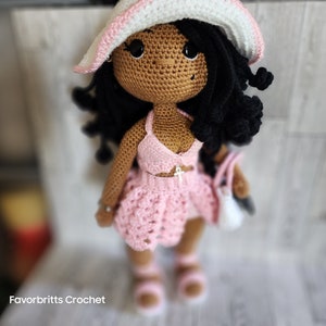 May include: Crocheted doll with brown skin, black hair, and a pink outfit. The doll is wearing a pink crochet hat, a pink crochet top, and a pink crochet skirt. The doll is holding a white crochet bag.