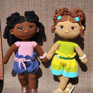 May include: Two crocheted dolls with brown skin, one wearing a pink and blue outfit and the other wearing a green and yellow outfit. Both dolls have brown hair and are holding hands.