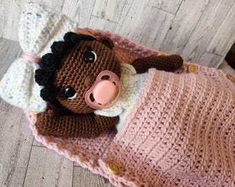 May include: A crocheted doll with brown skin, black hair, and a pink pacifier is lying in a pink crocheted sleeping bag with buttons.