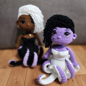 May include: Two crocheted dolls, one with white hair and a black dress, the other with black hair and a white dress. Both dolls have purple accents.