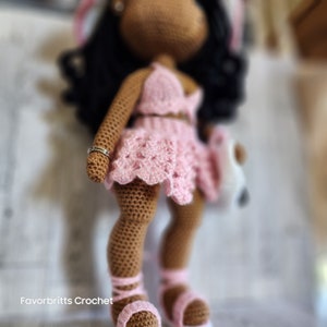 May include: A crocheted doll with brown skin, black hair, and a pink crocheted dress. The doll is wearing a silver ring on its finger and has a white crocheted bag in its hand.