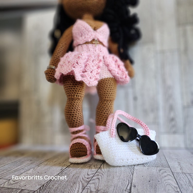 May include: A crocheted doll wearing a pink dress and sandals, holding a white crocheted bag with black heart-shaped sunglasses.