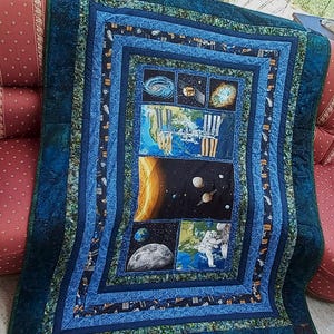 Op de afbeelding: Een handgemaakte quilt met een ruimtethema. De quilt heeft een centraal paneel met afbeeldingen van planeten, astronauten en de zon, omgeven door blauwe en groene patronen. De quilt is gemaakt van stof.
