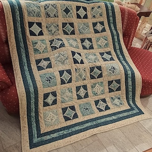 Könnte beinhalten: Eine handgefertigte Steppdecke mit einem sich wiederholenden Muster aus Quadraten und Diamanten. Die Steppdecke hat einen cremefarbenen Hintergrund mit türkisfarbenen und marineblauen Akzenten. Der Rand hat ein türkisfarbenes Streifendesign.