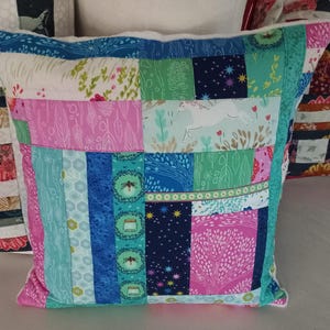 Può includere: Un cuscino patchwork con sfondo bianco e quadrati di tessuto di vari colori. I quadrati presentano diversi motivi, tra cui disegni floreali, geometrici e astratti. Il cuscino ha un bordo bianco e un bordo blu chiaro.