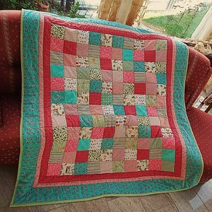 Op de afbeelding: Een patchwork quilt met een levendige mix van kleuren en patronen. De quilt heeft een vierkant ontwerp met verschillende stoffen vierkanten in roze, turquoise en groen. Het heeft een turquoise bloemenrand en een rode binnenrand.