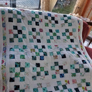 Könnte beinhalten: Eine weiße Steppdecke mit einem Schachbrettmuster aus verschiedenen Blautönen, Grüntönen und weißen Stoffquadraten. Die Steppdecke ist gefaltet und auf einem Sofa ausgestellt.