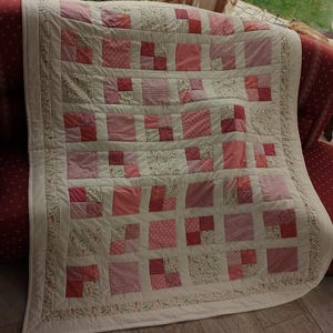 Op de afbeelding: Een handgemaakte quilt met een patchwork-ontwerp in roze en witte tinten. De quilt heeft vierkante blokken met verschillende patronen, waaronder bloemenprints en stippen, omzoomd door een bloemenrand. De quilt is over een rode bank gedrapeerd.