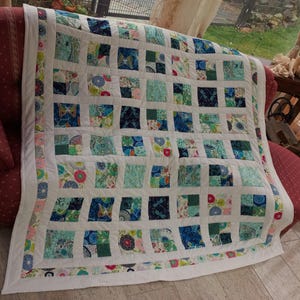 Könnte beinhalten: Eine weiße Steppdecke mit Patchwork-Muster, das aus Quadraten mit verschiedenen blauen, grünen und rosa Blumenmustern besteht. Die Steppdecke hat einen weißen Rand mit einem dünnen blauen Besatz.