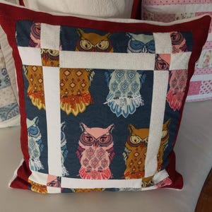 Op de afbeelding: Een rood en wit kussen met een blauwe achtergrond met een patchwork-ontwerp van uilen met verschillende patronen. De uilen zijn in verschillende kleuren, waaronder roze, geel en blauw.