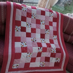 Könnte beinhalten: Eine rote und weiße Patchwork-Decke mit Blumenmuster. Die Decke besteht aus Quadraten aus verschiedenen Stoffen und ist mit einem weißen Rand eingefasst.