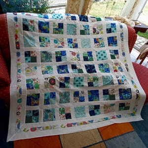 Könnte beinhalten: Eine Patchwork-Decke mit einem Gitter aus quadratischen Stoffblöcken in verschiedenen Blau-, Grün- und Weißtönen. Die Decke ist von einem weißen Stoffrahmen und einem dünnen äußeren Streifen aus gemustertem Stoff mit kleinen Blumenmotiven umrandet.