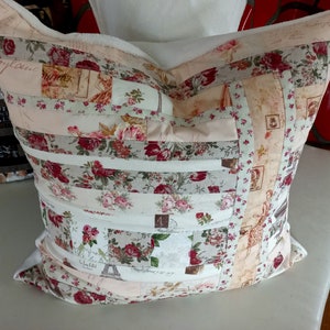 Può includere: Un cuscino decorativo con un design a patchwork che presenta motivi floreali in tonalità di rosa, rosso e bianco. Il cuscino è realizzato con ritagli di tessuto cuciti insieme in un motivo a strisce. Il cuscino ha uno sfondo bianco ed è accentuato da tessuto beige e marrone.