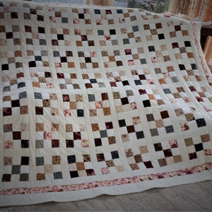 Könnte beinhalten: Eine weiße Steppdecke mit Patchwork-Muster, das aus Quadraten in verschiedenen Brauntönen, Beige, Grau und Rot besteht. Die Steppdecke hat einen dekorativen Rand mit einem floralen Muster in Rosa- und Rottönen.