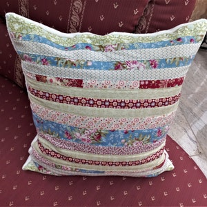 Può includere: Un cuscino decorativo con un design a patchwork che presenta strisce di vari tessuti floreali in tonalità di blu, rosa, verde e rosso. Il cuscino ha un bordo bianco ed è appoggiato su un tessuto color borgogna.