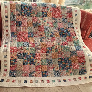 Può includere: Una trapunta patchwork con bordo bianco e una varietà di motivi floreali e paisley colorati. La trapunta è drappeggiata su una poltrona rossa. I quadrati della trapunta presentano sfumature di rosso, blu, verde e giallo.