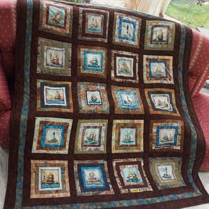 Könnte beinhalten: Eine braune und blaue Quiltdecke mit nautischem Thema. Die Decke besteht aus Quadraten mit verschiedenen Bildern von Schiffen und Leuchttürmen. Die Decke ist mit einem braunen und blauen Rand eingefasst.
