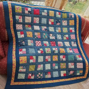 Könnte beinhalten: Eine handgefertigte Patchwork-Decke mit blauem Denim-Hintergrund und bunten quadratischen Flicken. Die Quadrate zeigen florale und gemusterte Stoffe in Rot, Gelb, Rosa und Grün. Die Decke hat einen gelben Blumenrand.
