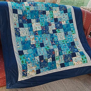 Könnte beinhalten: Ein Patchwork-Quilt mit maritimem Thema, mit Quadraten aus verschiedenen blauen Stoffen mit Bildern von Segelbooten, Meeresbewohnern und Leuchttürmen. Der Quilt hat einen weißen Rand und eine dunkelblaue Rückseite und ist ein dekorativer Artikel.