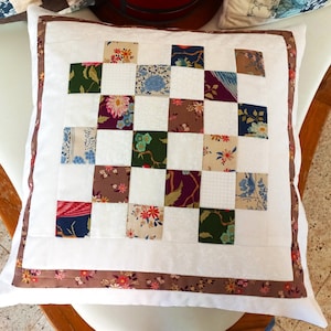 Peut inclure: Un oreiller blanc avec une bordure brune et un motif patchwork de différents motifs floraux et géométriques dans des tons de rouge, vert, bleu et marron.