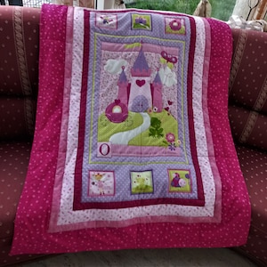 Könnte beinhalten: Eine rosafarbene und violette gesteppte Babydecke mit einem Prinzessinnen-Schloss-Design. Die Decke zeigt ein großes Schloss mit einem Herzen auf der Vorderseite, eine Kutsche, einen Frosch und den Buchstaben O. Der Text auf der Decke lautet "Es war einmal eine kleine alte Prinzessin."