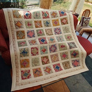Pode incluir: Uma colcha de retalhos retangular com uma grade de blocos quadrados, cada um contendo uma forma de diamante central em vários padrões florais e geométricos. A colcha tem uma borda cor de creme e uma almofada quadrada a condizer.