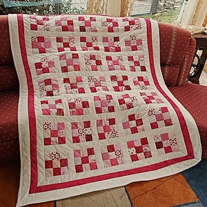 Può includere: Una trapunta patchwork caratterizzata da una griglia di blocchi a nove toppe in varie tonalità di rosa, rosso e bianco. La trapunta è incorniciata da un bordo interno rosa a tinta unita e un bordo esterno bianco più ampio, con cuciture visibili.