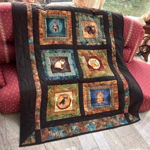 Könnte beinhalten: Eine braune und schwarze Steppdecke mit Patchwork-Muster, das verschiedene Katzen in verschiedenen Posen zeigt. Die Steppdecke ist aus Stoff mit verschiedenen Farben und Mustern gefertigt, darunter Braun, Schwarz, Weiß und Blau. Die Steppdecke wird auf einem Sofa ausgestellt.