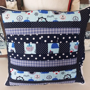 Può includere: Un cuscino patchwork blu e bianco a tema nautico. Il cuscino presenta balene, ancore e navi. Il tessuto è un mix di pois, quadri e colori solidi.