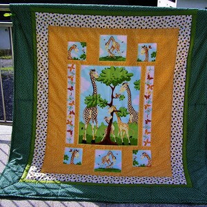 Könnte beinhalten: Eine handgefertigte Steppdecke mit Giraffenmotiv. Die zentrale Tafel zeigt Giraffen unter einem Baum, umgeben von kleineren quadratischen Tafeln mit Giraffenillustrationen. Die Steppdecke hat ein gelbes, weißes und grünes Farbschema mit einem gepunkteten Rand.