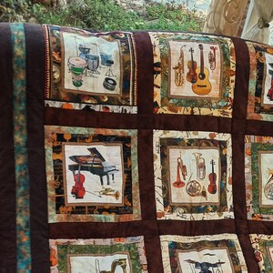 Könnte beinhalten: Ein brauner gesteppter Stoff mit einem Patchwork-Muster, das verschiedene Musikinstrumente zeigt. Die Quadrate sind mit Bildern eines Schlagzeugs, eines Klaviers, eines Cellos, einer Trompete, einer Gitarre und einer Geige verziert. Der Stoff hat einen braunen Hintergrund mit einem grünen und blauen Rand.