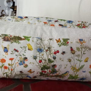 Könnte beinhalten: Weiße Stoffaufbewahrungstasche mit einem Blumendruck mit Feen, Schmetterlingen und Blumen. Die Tasche hat einen weißen Spitzenbesatz und einen Reißverschluss.