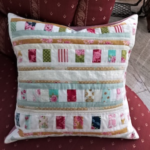 Op de afbeelding: Een wit kussen met een patchwork-ontwerp met verschillende bloemen- en geometrische patronen in tinten roze, groen, blauw en goud. Het kussen is gemaakt van verschillende stoffen, waaronder kant en strepen.