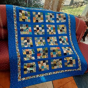 Könnte beinhalten: Eine Patchwork-Decke mit sechzehn quadratischen Blöcken, die jeweils aus kleineren, bunten Stoffquadraten mit verschiedenen Mustern bestehen. Die Blöcke sind durch einfarbige blaue Streifen getrennt und von einer blauen Bordüre mit Affenmuster umrahmt.