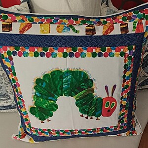 Könnte beinhalten: Ein dekoratives Kissen mit einem farbenfrohen Design. Es zeigt eine große grüne Raupe mit rotem Kopf und lila Antennen. Der Rand ist mit bunten Punkten und Lebensmittelillustrationen verziert.