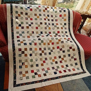 May include: A patchwork quilt featuring a grid of small, multi-colored fabric squares in shades of red, blue, yellow, and green, separated by white sashing. The quilt is finished with a dark floral border and a wide, solid white outer border.