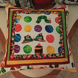 Può includere: Cuscino quadrato decorativo con un design colorato raffigurante un bruco, una farfalla e vari alimenti. Il cuscino ha uno sfondo bianco con grandi pois colorati e un bordo a fantasia. Il design ricorda un'illustrazione di un libro per bambini.