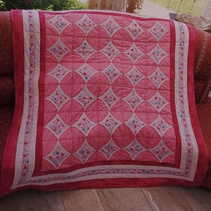 Könnte beinhalten: Eine rechteckige Steppdecke mit einem sich wiederholenden Muster aus rosa und weißen Rautenformen. Die Mitte zeigt ein Gitter aus rosa Stoffquadraten mit weißen, geschwungenen Kanten, akzentuiert durch kleine florale Motive in Blau und Rosa. Ein breiter rosa Rand rahmt die Decke ein.