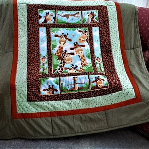 Könnte beinhalten: Eine grüne, braune und orangefarbene Patchwork-Decke mit einem zentralen Feld, das eine Gruppe von Giraffen mit verspielten Gesichtsausdrücken zeigt. Die Giraffen sind von einem grünen und blauen Himmel mit weißen Wolken umgeben. Die Decke ist mit einem braunen Giraffenstoff eingefasst.
