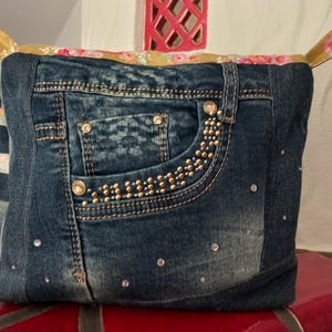 Könnte beinhalten: Ein Aufbewahrungskorb aus Jeansstoff mit einer Tasche, die mit goldfarbenen Nieten und Strasssteinen verziert ist. Der obere Rand und die Griffe sind mit floralem Stoff versehen. Der Korb ist wie eine Jeans gestaltet.