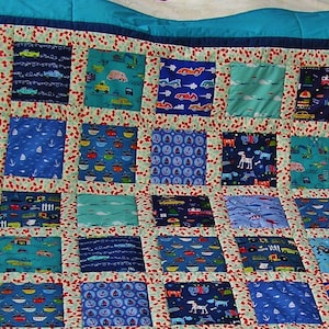 Op de afbeelding: Een blauwe en witte patchworkdeken met verschillende ontwerpen, waaronder auto's, boten en dieren. De deken is gemaakt van verschillende stoffen en heeft een lichtgroene achtergrond.