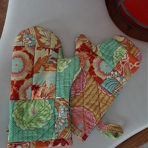 Könnte beinhalten: Ein Paar gesteppte Ofenhandschuhe und ein Topflappen mit Patchwork-Design. Die Handschuhe und der Topflappen weisen Blumen- und Blättermuster in Orange-, Grün-, Gelb- und Rosatönen auf. Die Handschuhe sind etwa 33 cm lang.