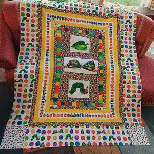 Könnte beinhalten: Eine rechteckige Steppdecke mit weißem Hintergrund und einem wiederkehrenden Muster aus bunten Kreisen, die Illustrationen von Früchten, Kuchen und Raupen enthalten. Die Mitte zeigt drei weiße Felder mit einem grünen Blatt, einem Schmetterling und einer Raupe.